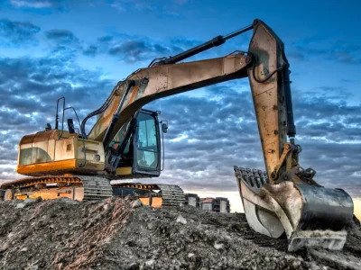 Yellow tracked excavator with its bucket lowered on a graded dirt pad under a cloudy sky